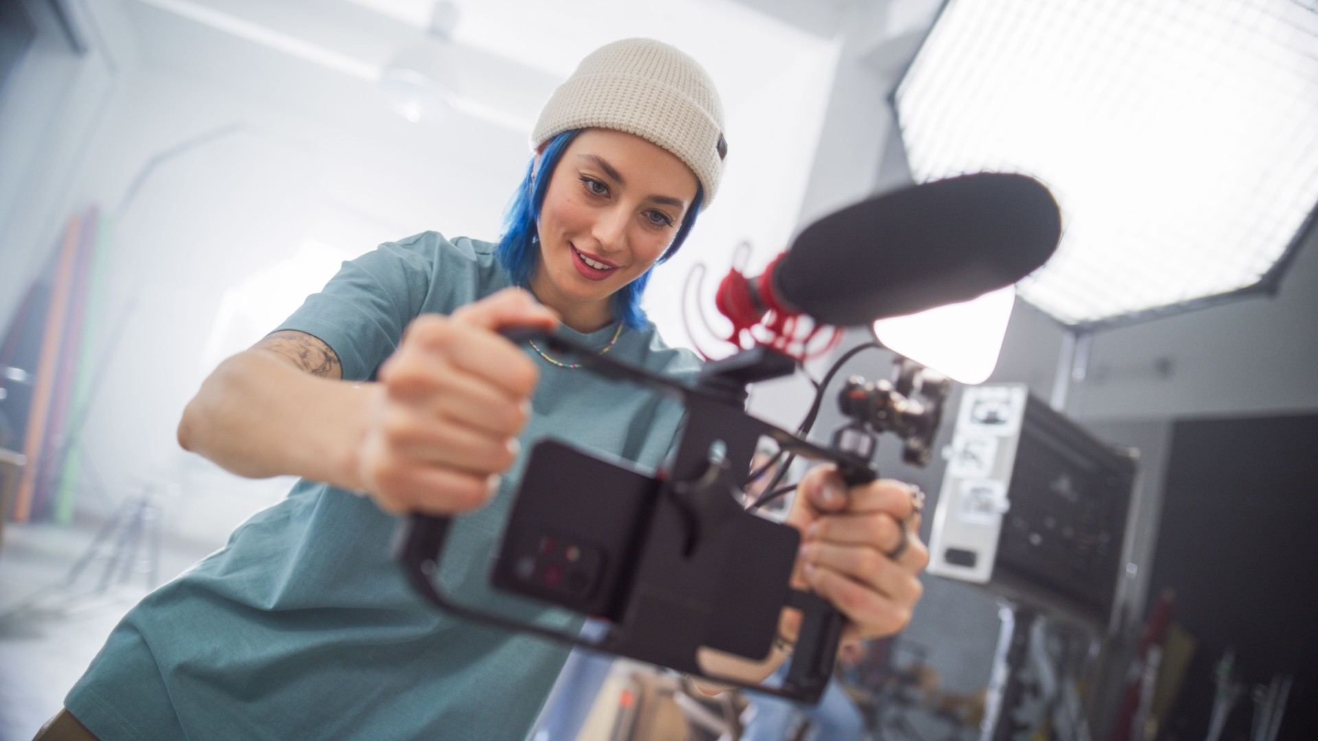 Young Caucasian Female Cinematographer With Blue Hair Adjusts Camera Equipment In Modern Studio, Focusing On Creative Video Production For Advertising, Showcasing Teamwork And Artistic Skill.