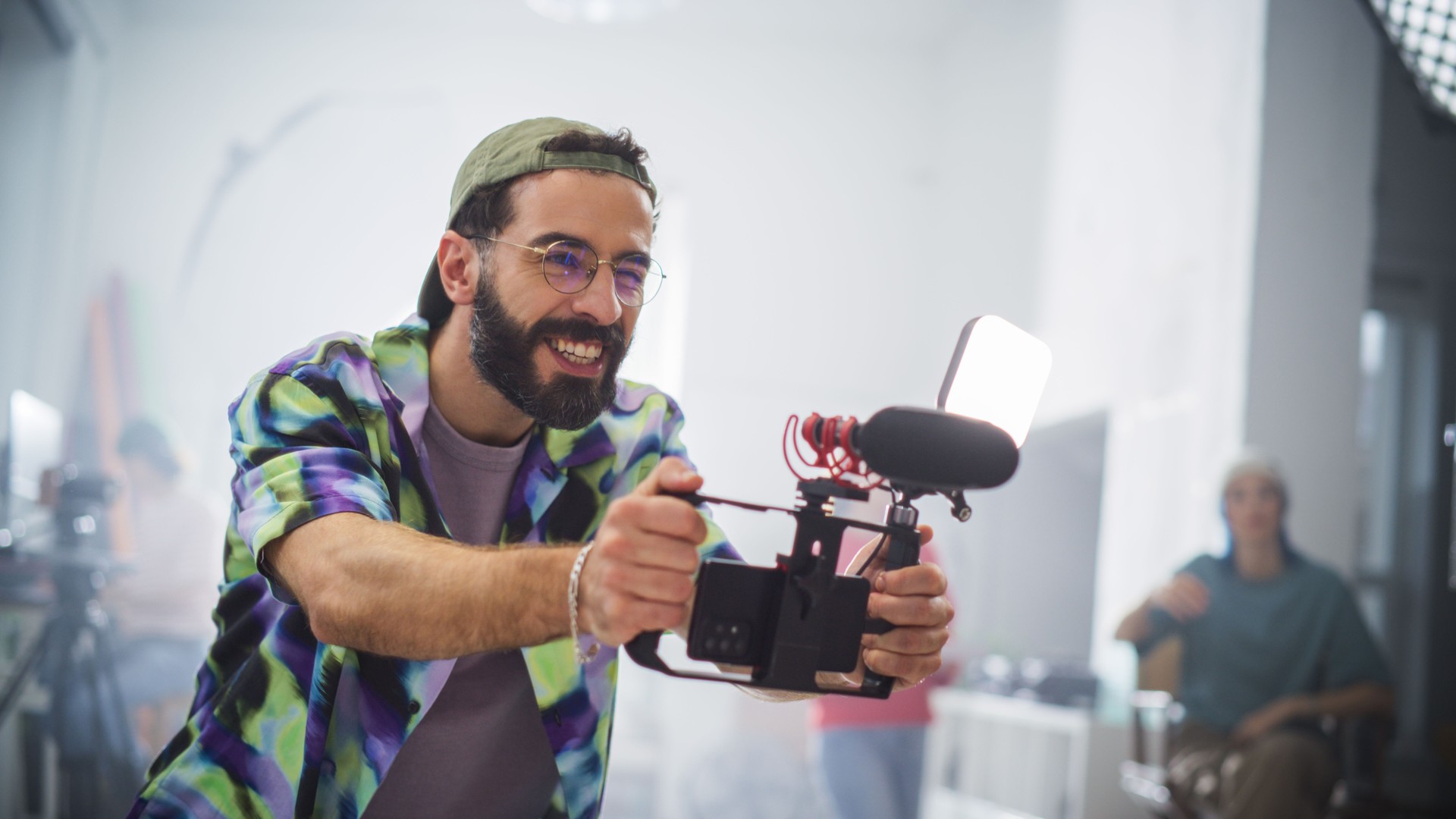 Young Caucasian Male Videographer With Beard, Wearing Colorful Shirt And Cap, Joyfully Operates Camera Equipment In A Well-lit Studio, Surrounded By Creative Team Members Engaged In Film Production.